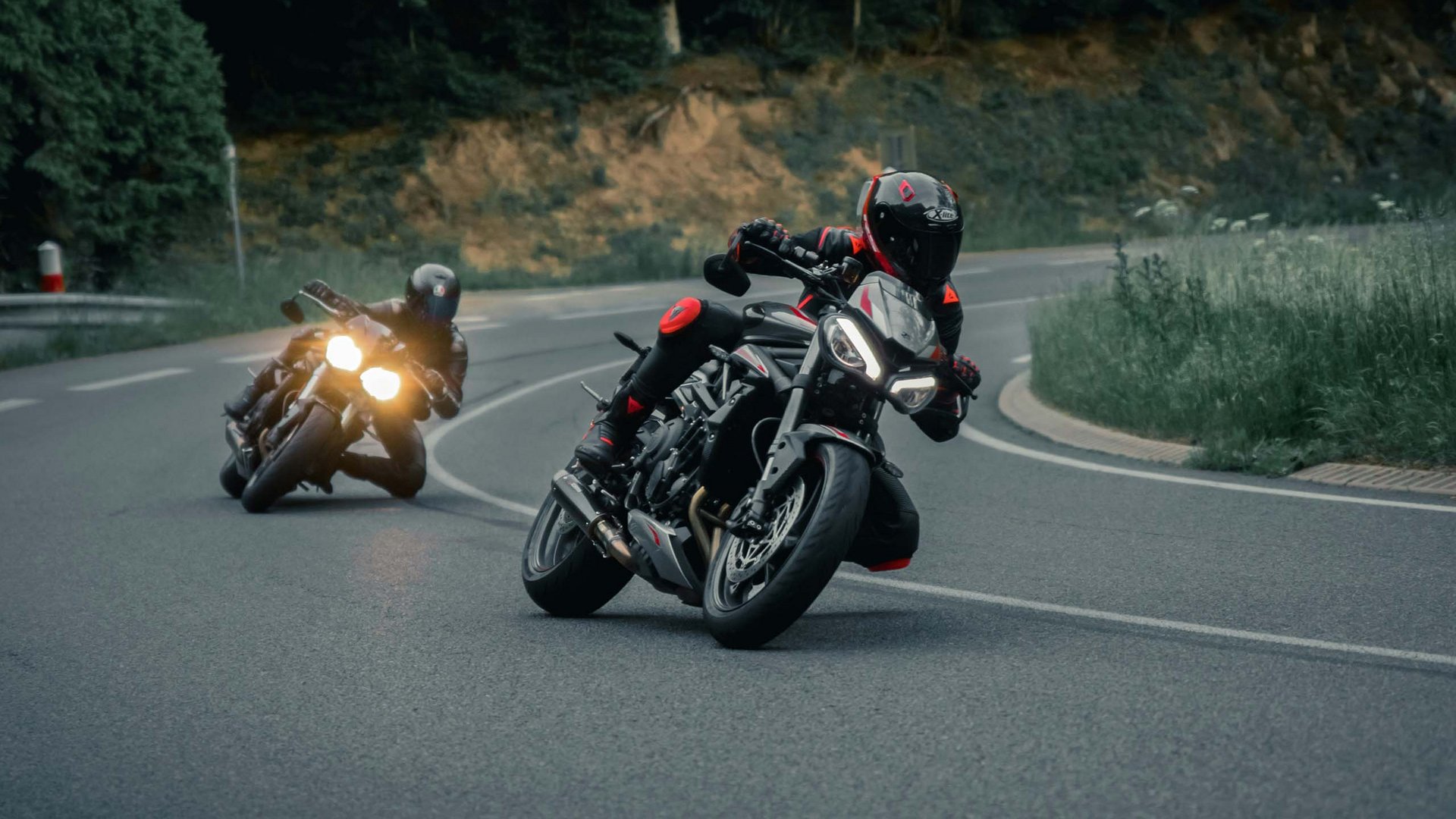 Your motorcycle hotel in the Bavarian Forest Two motorcyclists in black gear riding on a winding road through forest